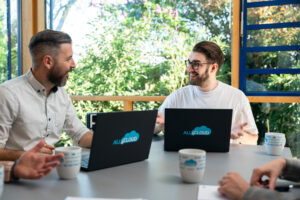 Zwei Männer sitzen mit Laptops an einem Tisch, lächeln und unterhalten sich während eines Meetings über SAP Cloud ERP Erweiterungen. Sonnenlicht fällt durch große Fenster, draußen ist Grün, auf dem Tisch liegen Kaffeetassen und Hände anderer Personen.