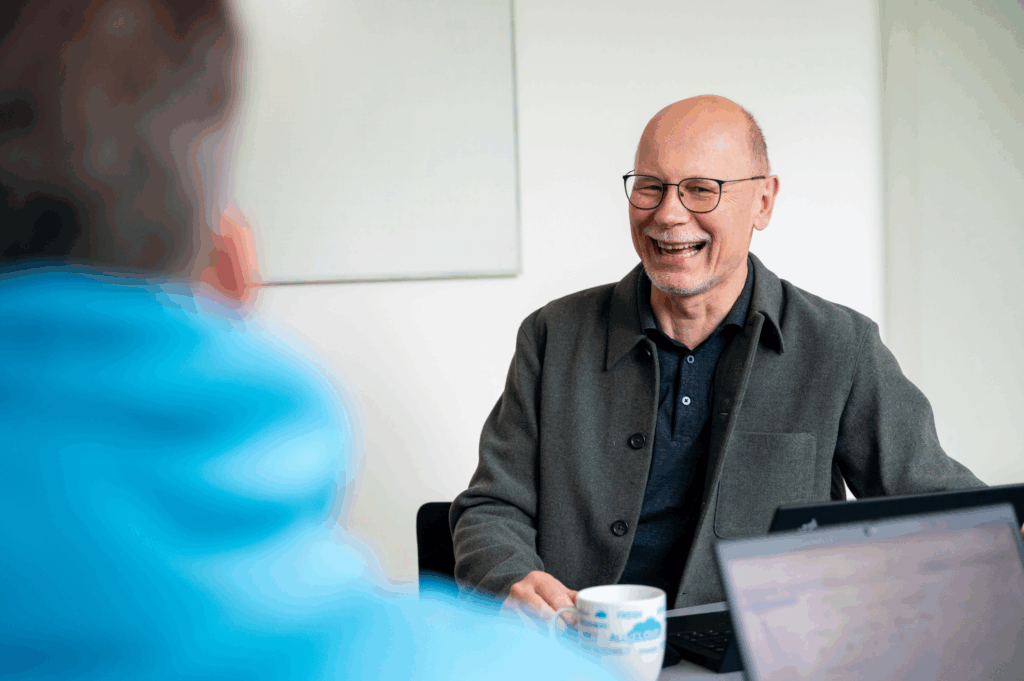 Ein lächelnder, glatzköpfiger Mann mit Brille sitzt an einem Tisch mit Laptop und Kaffeetasse und diskutiert mit einer anderen Person in einem blauen Kapuzenpulli, die im Vordergrund unscharf ist, über SAP Cloud ERP für Stadtwerke und Versorger.