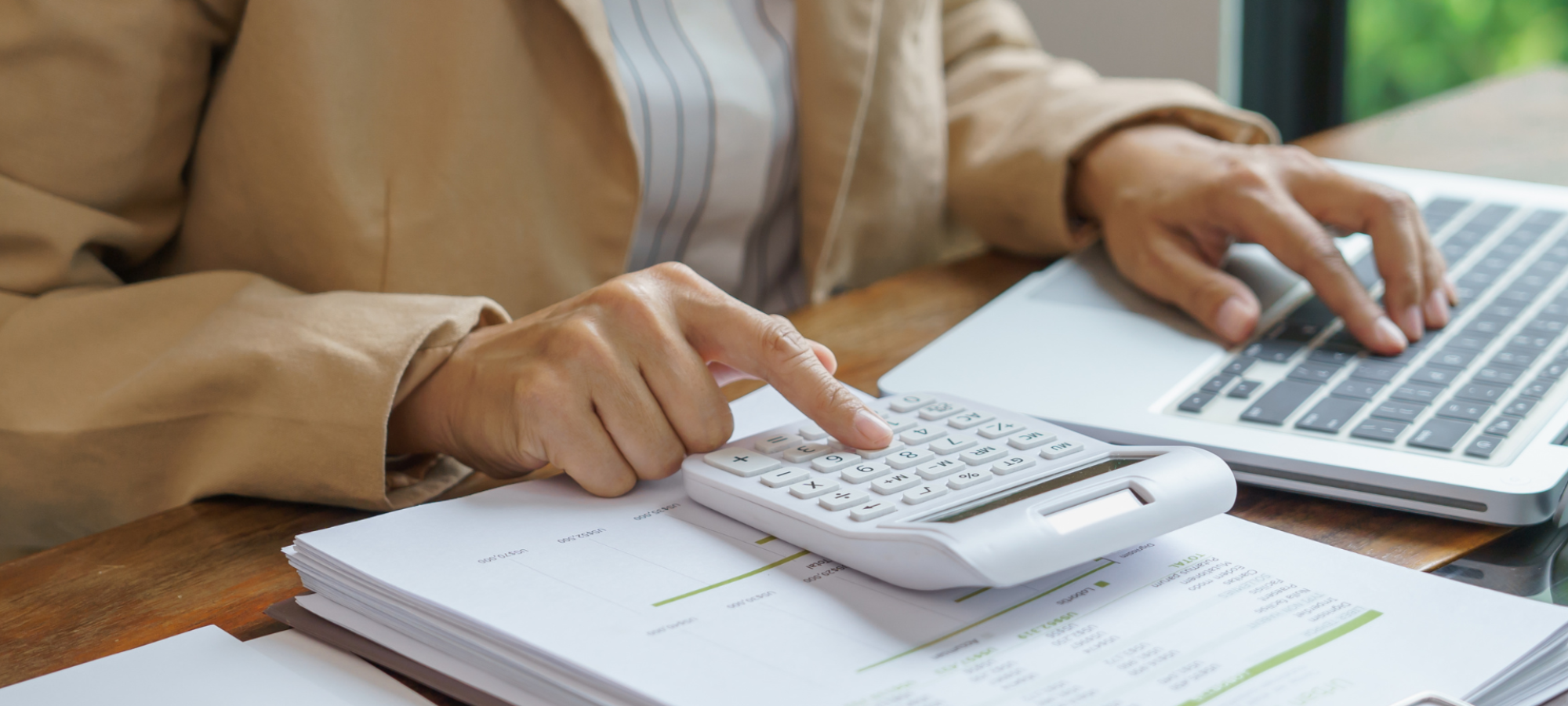 Eine Person in brauner Jacke arbeitet mit einem Taschenrechner und einem Laptop mit SAP Cloud ERP an einem Schreibtisch, vor dem Dokumente und Diagramme ausgebreitet sind, um Fast Close für einen effizienten Abschluss zu beschleunigen.