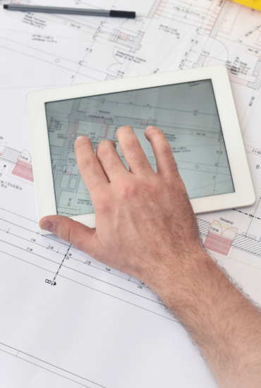 Eine Person mit weißem Schutzhelm arbeitet mit einem Tablet, auf dem Architekturpläne angezeigt werden, umgeben von Papierplänen, einem Smartphone und Zeichenwerkzeugen auf einem Schreibtisch - eine Demonstration der Effizienz für technische Dienstleister mit SAP Cloud ERP.