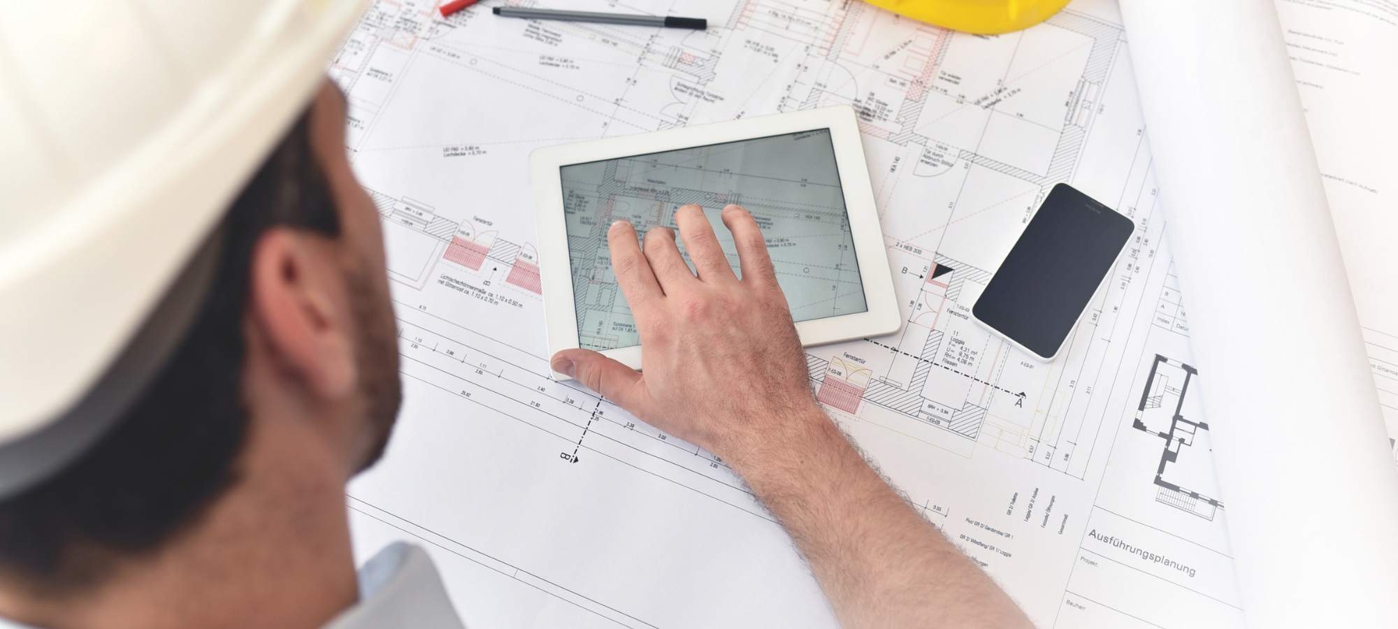 Eine Person mit weißem Schutzhelm arbeitet mit einem Tablet, auf dem Architekturpläne angezeigt werden, umgeben von Papierplänen, einem Smartphone und Zeichenwerkzeugen auf einem Schreibtisch - eine Demonstration der Effizienz für technische Dienstleister mit SAP Cloud ERP.