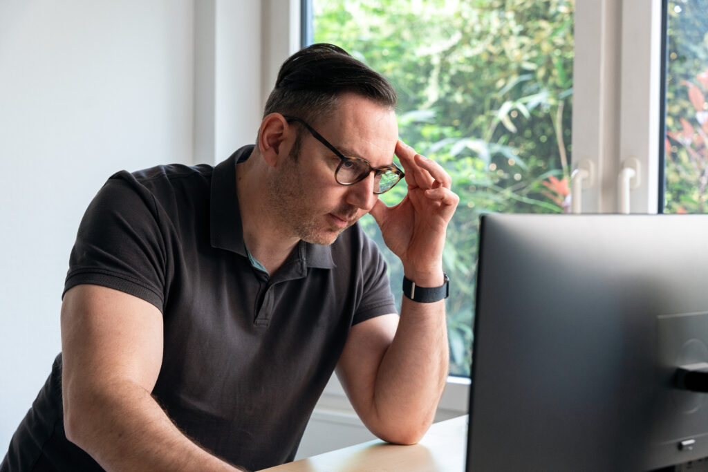 A man wearing glasses and a black polo shirt sits at a desk, looking thoughtfully at a computer monitor. There are large windows behind him with green foliage visible outside.