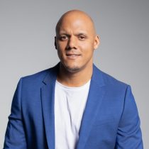 Ein Mann mit rasiertem Kopf trägt einen blauen Blazer über einem weißen Hemd und lächelt leicht. Der Hintergrund ist einfarbig und hellgrau.