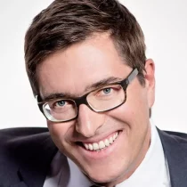 Ein Mann mit kurzen braunen Haaren und Brille lächelt in die Kamera. Er trägt einen dunklen Anzug, ein weißes Hemd und eine Krawatte und steht vor einem schlichten weißen Hintergrund.