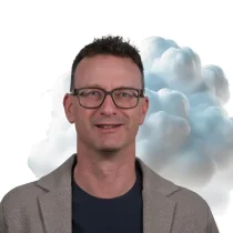 Ein Mann mit kurzen Locken und Brille, der eine Jacke über einem dunklen Hemd trägt, steht vor einem weißen Hintergrund, hinter ihm ist eine wolkenähnliche Formation zu sehen.