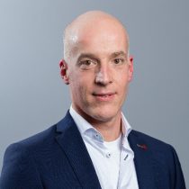 Ein kahlköpfiger Mann in einem marineblauen Anzug und einem weißen Hemd posiert vor einem schlichten grauen Hintergrund und blickt mit ausdruckslosem Gesicht direkt in die Kamera.