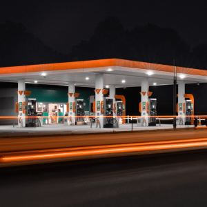 Eine hell erleuchtete Holidu-Tankstelle mit orangefarbenen und weißen Akzenten wird bei Nacht fotografiert. Im Bild sind mehrere leere Zapfsäulen zu sehen, und im Vordergrund huschen verschwommene Autoscheinwerfer vorbei.