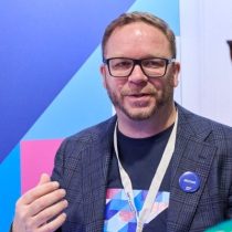 Ein Mann mit Brille und Bart, der einen blauen Blazer und ein Namensschild mit der Aufschrift „Michael“ trägt, steht vor einem bunten geometrischen Hintergrund und gestikuliert mit einer Hand.